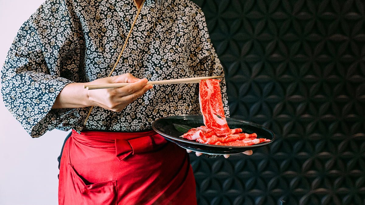 Woman wearing kimono holding rare slice Wagyu A5 beef by chopsticks for boiling in Sukiyaki hot pot.